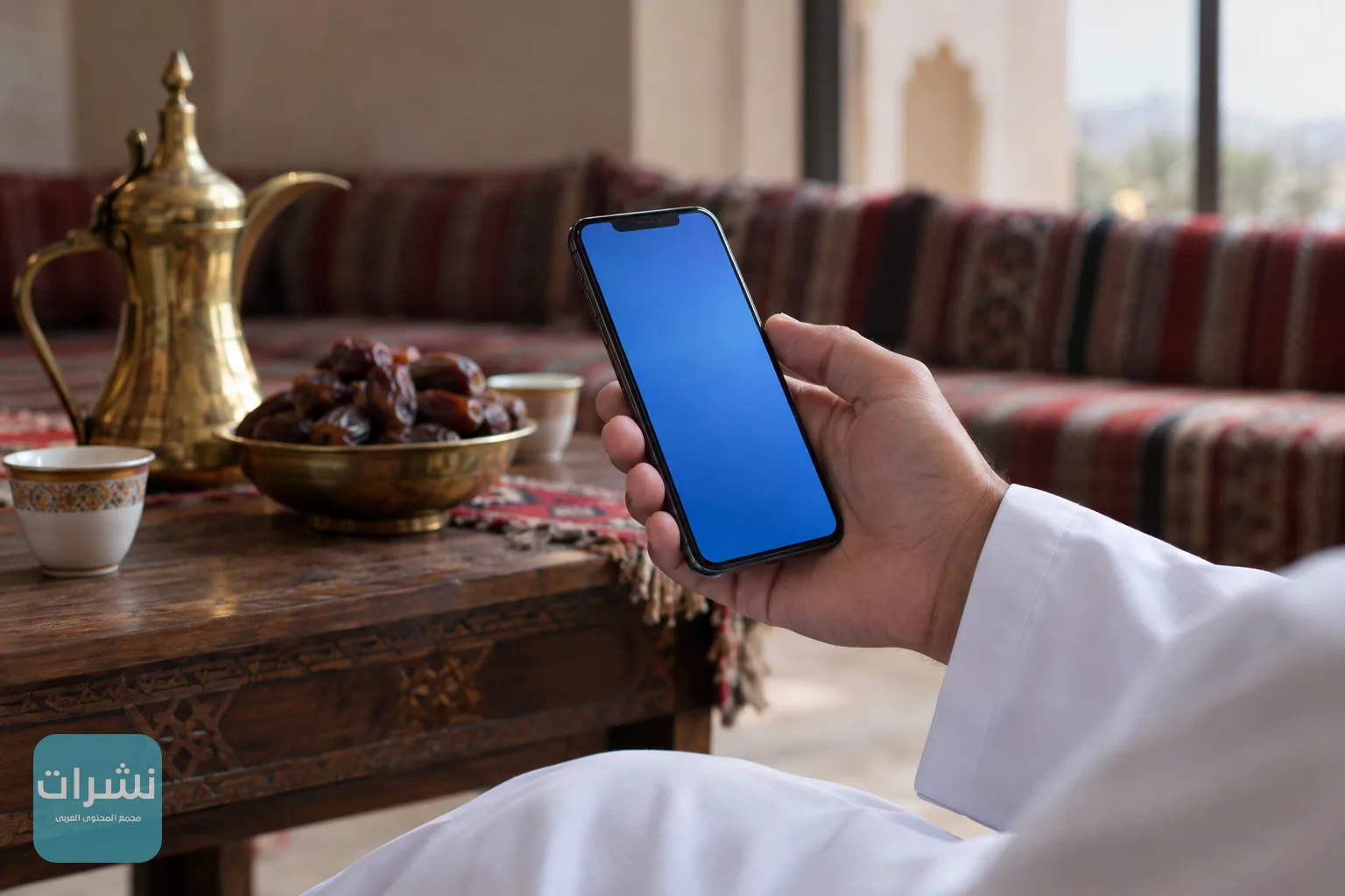 يد تمسك هاتفًا ذكيًا في مجلس عُماني تقليدي بجانب طبق من التمر