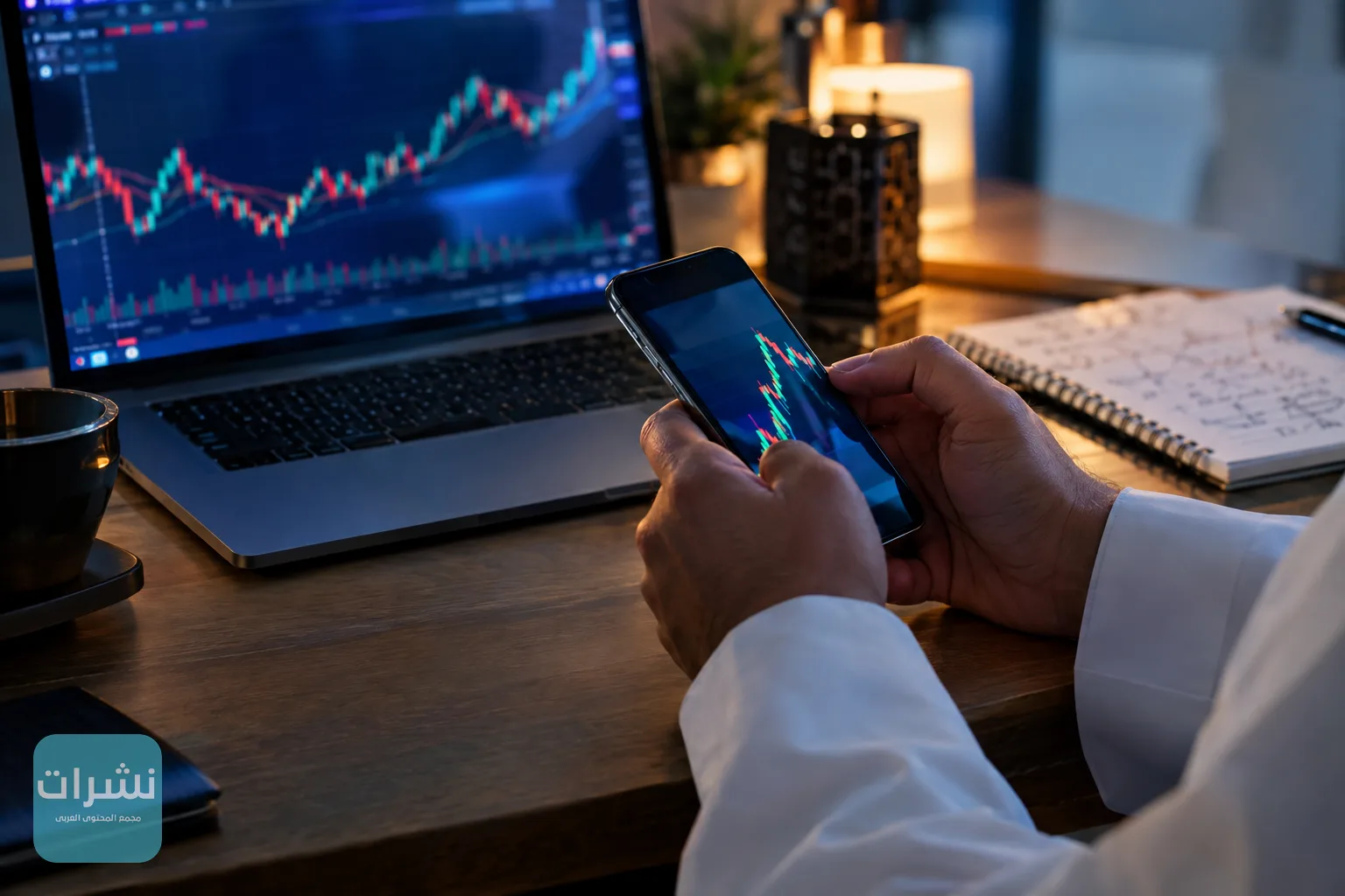 يد متداول تمسك هاتفًا أمام حاسوب تنعكس عليه مخططات أسعار غير مقروءة
