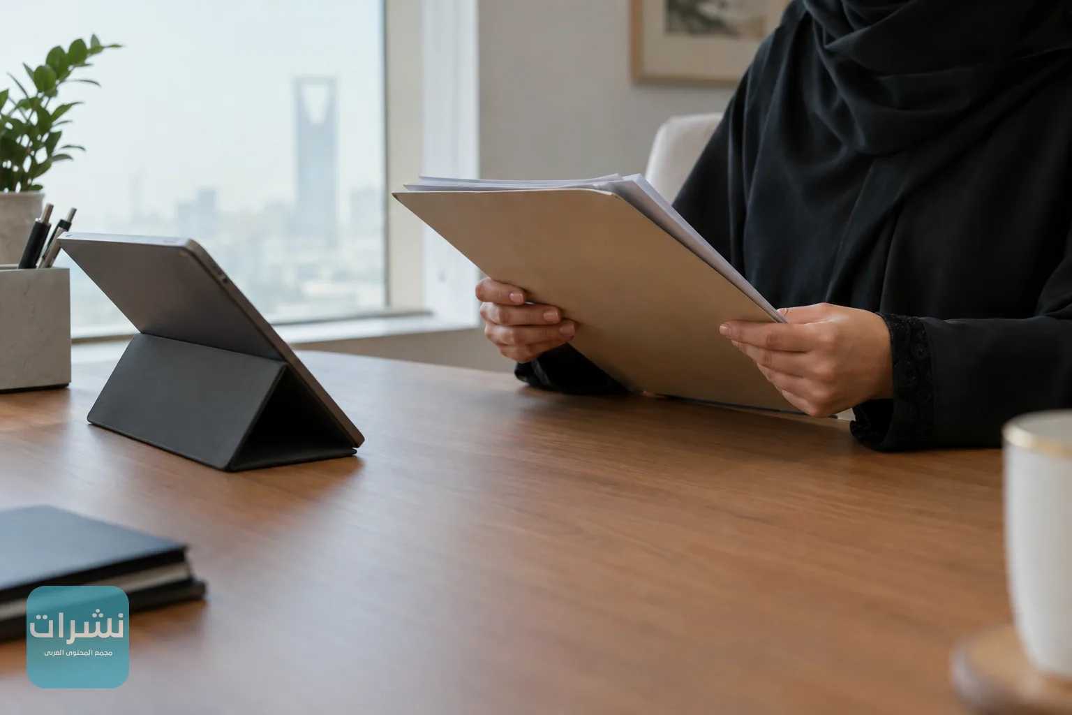 يد تحمل ملف أوراق بجانب جهاز لوحي على مكتب خشبي في مكتب صغير