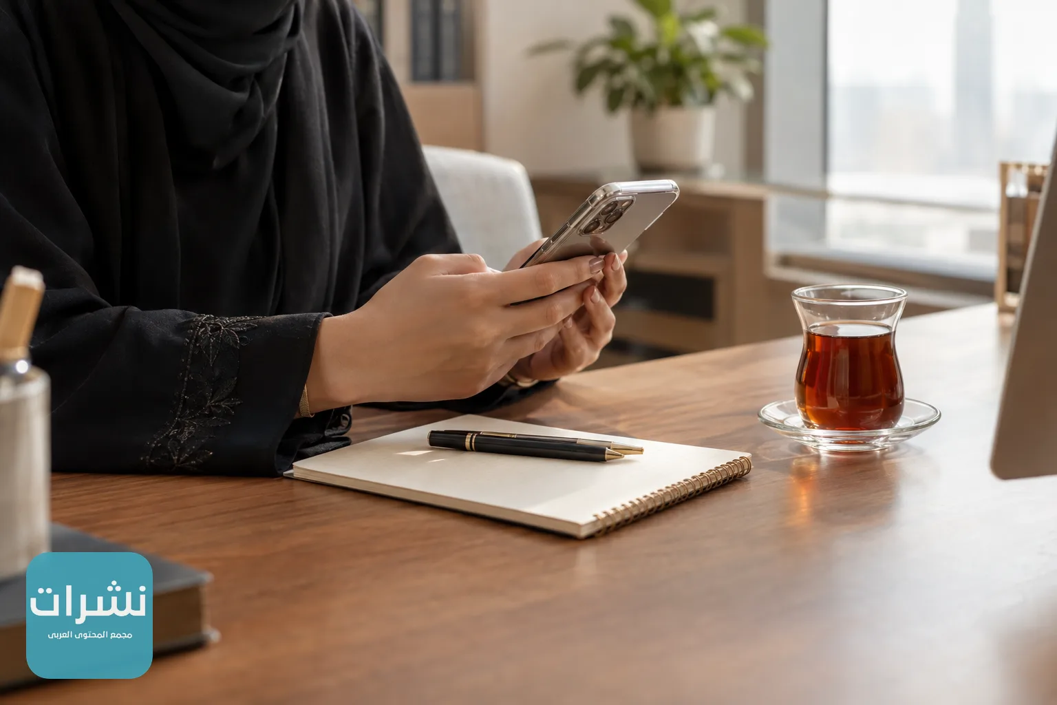 معلمة تمسك هاتفها أمام دفتر وأوراق على مكتب في مكان عمل هادئ